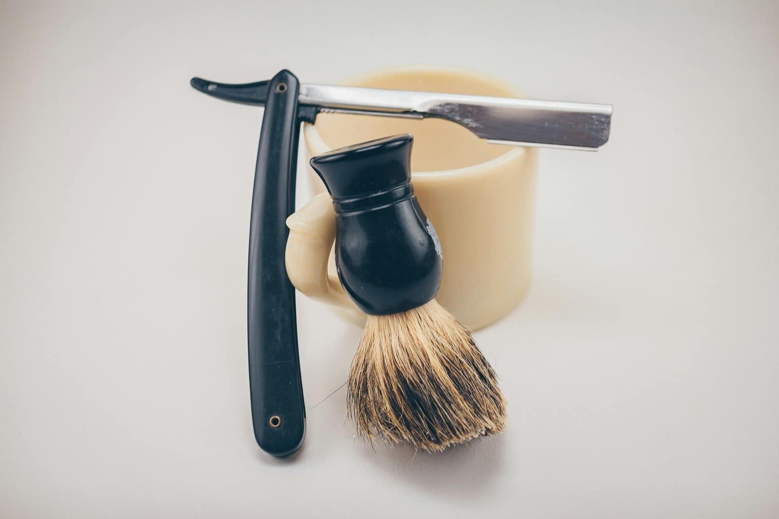 A black straight razor and a shaving brush resting against a cream shaving mug on a pale background